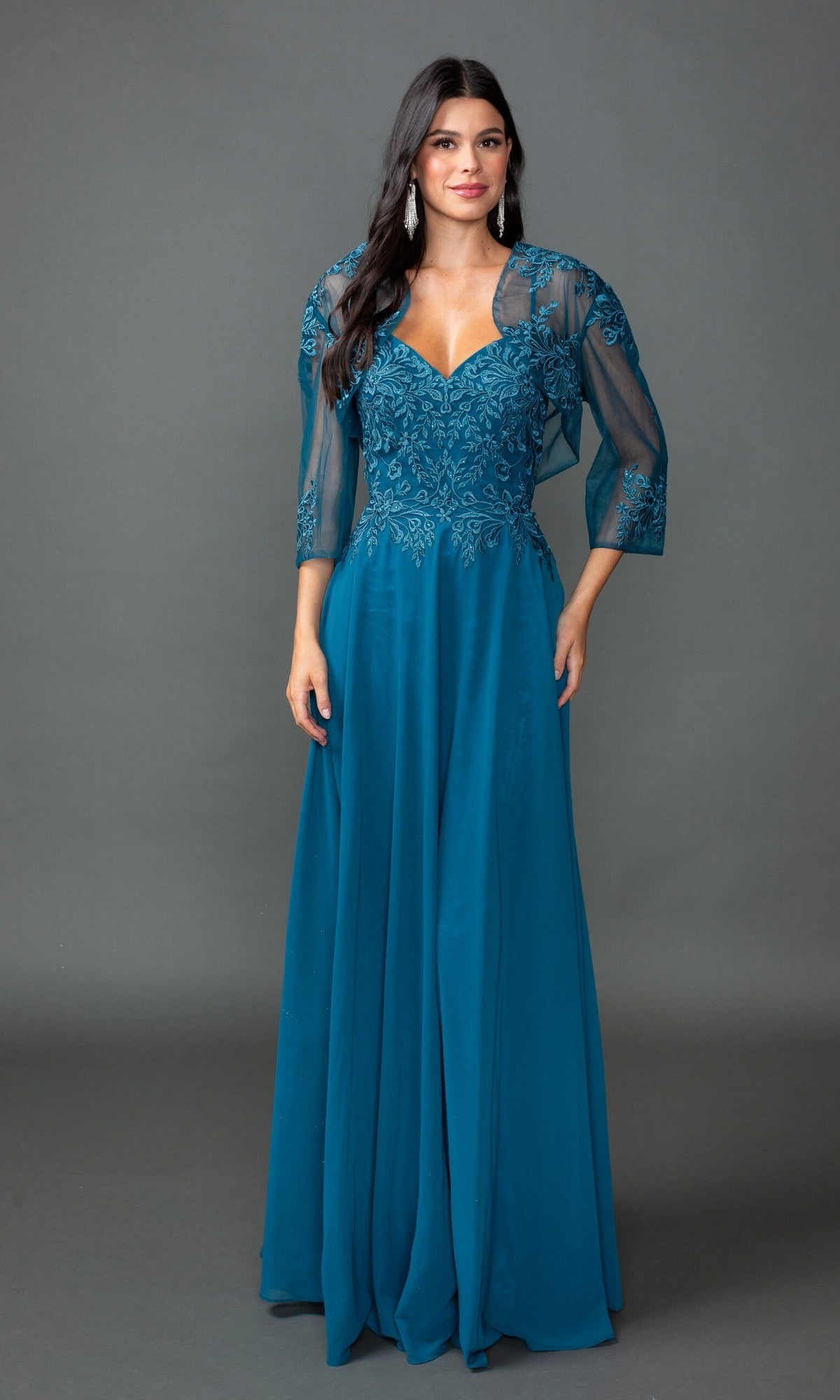 A woman models the Dancing Queen 4542 long prom dress in teal, featuring lace embroidery and a sheer matching shawl, against a plain gray backdrop.